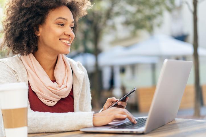 woman holding phone and looking at laptop outside, cup of coffee