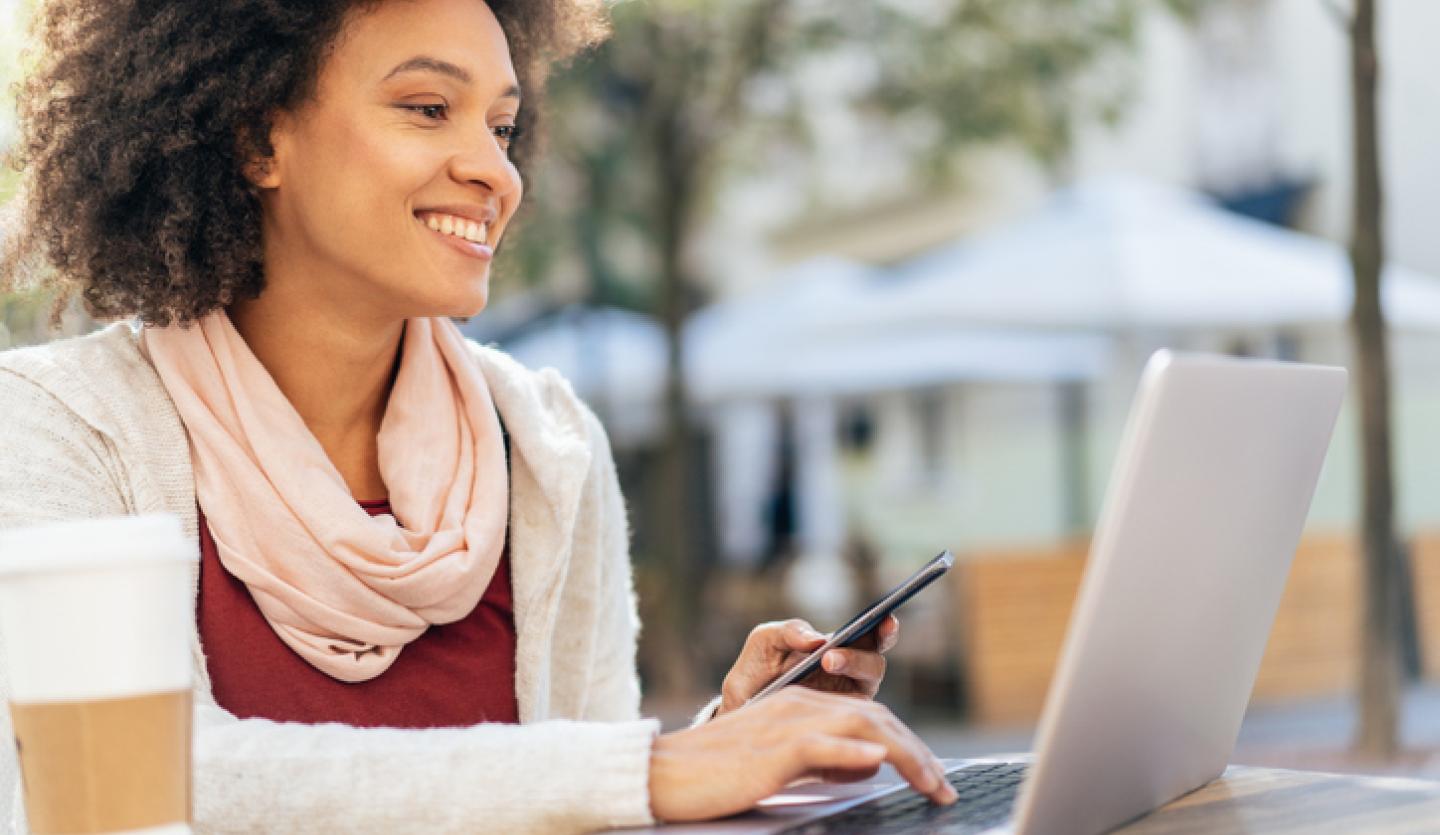 woman holding phone and looking at laptop outside, cup of coffee