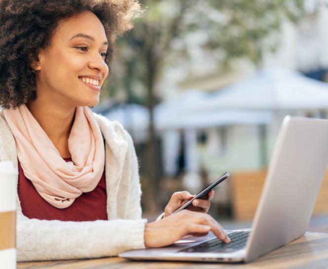 woman holding phone and looking at laptop outside, cup of coffee