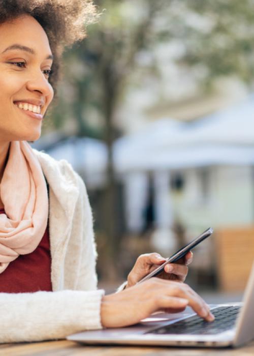 woman holding phone and looking at laptop outside, cup of coffee